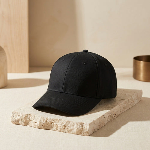 The Luxuel Laser Cap 272, a matte black six-panel cap with a topstitched brim, resting on a travertine plinth in warm daylight